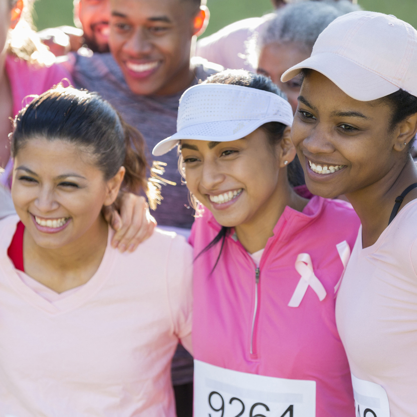 This is a high angle closeup of a group photo of men and women with their arms around each other. They are participants in a race for the cure of breast cancer.